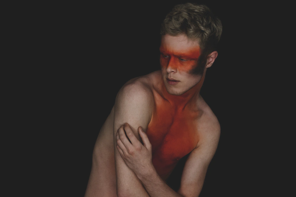 Portrait of a young man, bare-chested, against a black background, with a strip of orange light crossing his eyes like a mask